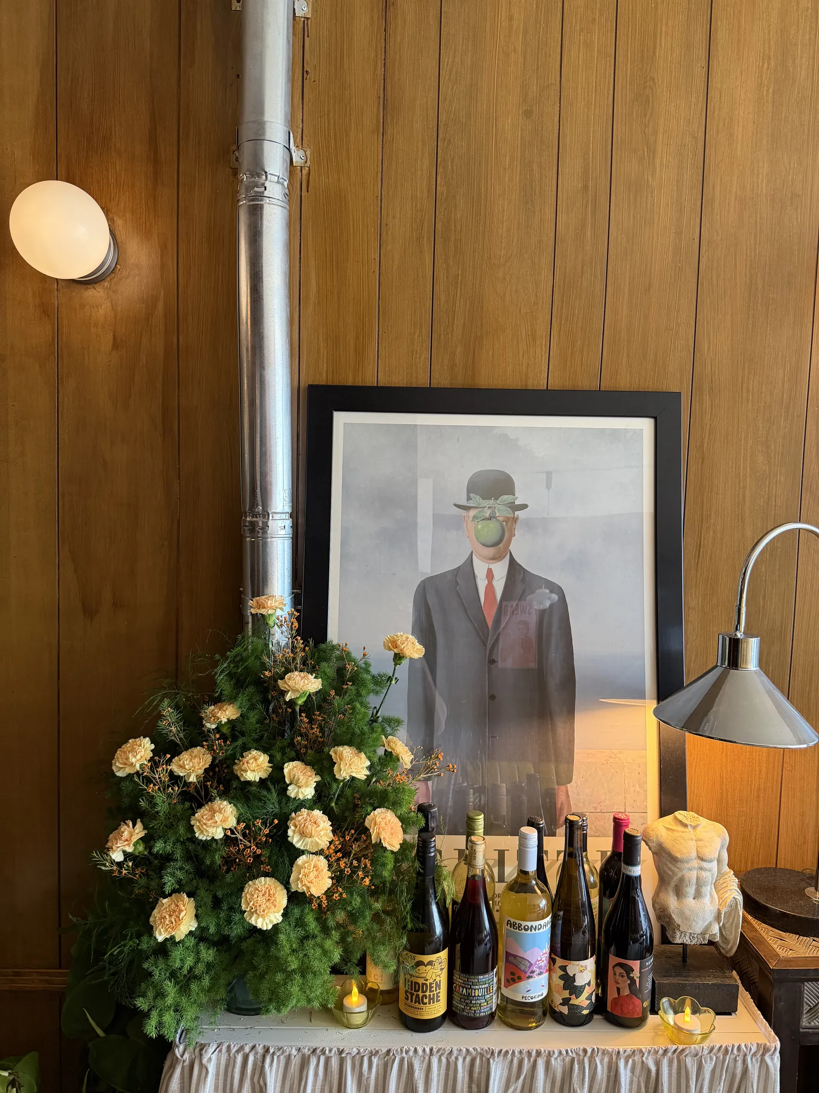 A corner of the dining room with yellow roses, wine bottles and a framed painting